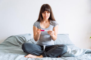 Mujer sentada en la cama con las piernas en cruzadas, mirando un test de embarazo y preguntándose: ¿porque no me quedo embarazada?