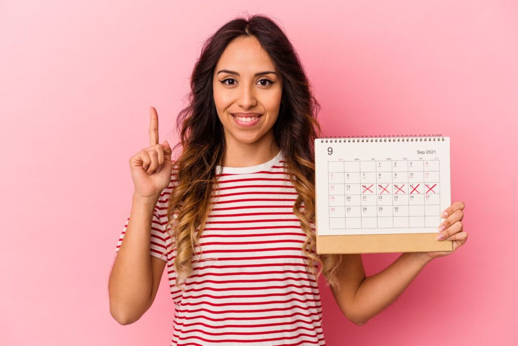 young mexican woman holding calendar isolated pink background showing number one with finger min